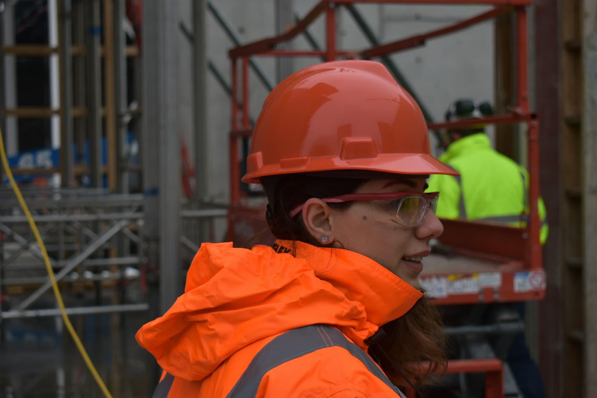 Construction worker in an orange hard hat, safety glasses and hi-vis jacket on site, with a scissor lift and scaffold behind